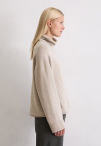 Beżowy sweter z golfem, wykonany z miękkiego materiału, o luźnym kroju, z prążkowanymi mankietami i subtelną fakturą, w połączeniu z ciemnymi spodniami.