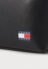 Čierna textúrovaná taška s gumovaným modro-bielo-červeným obdĺžnikovým logom s nápisom "TOMMY JEANS" na prednej strane.