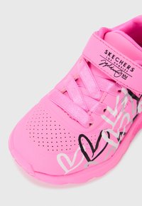 Scarpa da ginnastica rosa con una parte superiore sintetica liscia, chiusura con lacci, design perforato e grafiche a cuore bianche e nere.