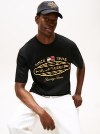 Svart t-skjorte med gull "HILFIGER Racing Team" logo, rund hals, kort ermer, kombinert med hvite bukser og en matchende caps.