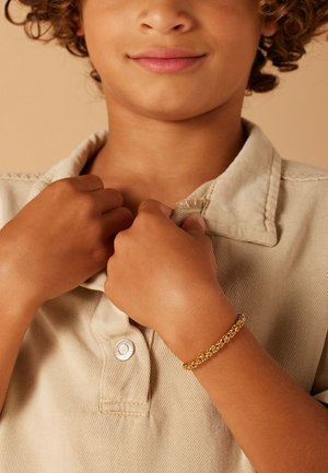Gouden armband om de pols van een kind, met een gekoppeld ontwerp. Het kind draagt een lichtbeige overhemd met kraag en zichtbare knoop- en stikdetails.