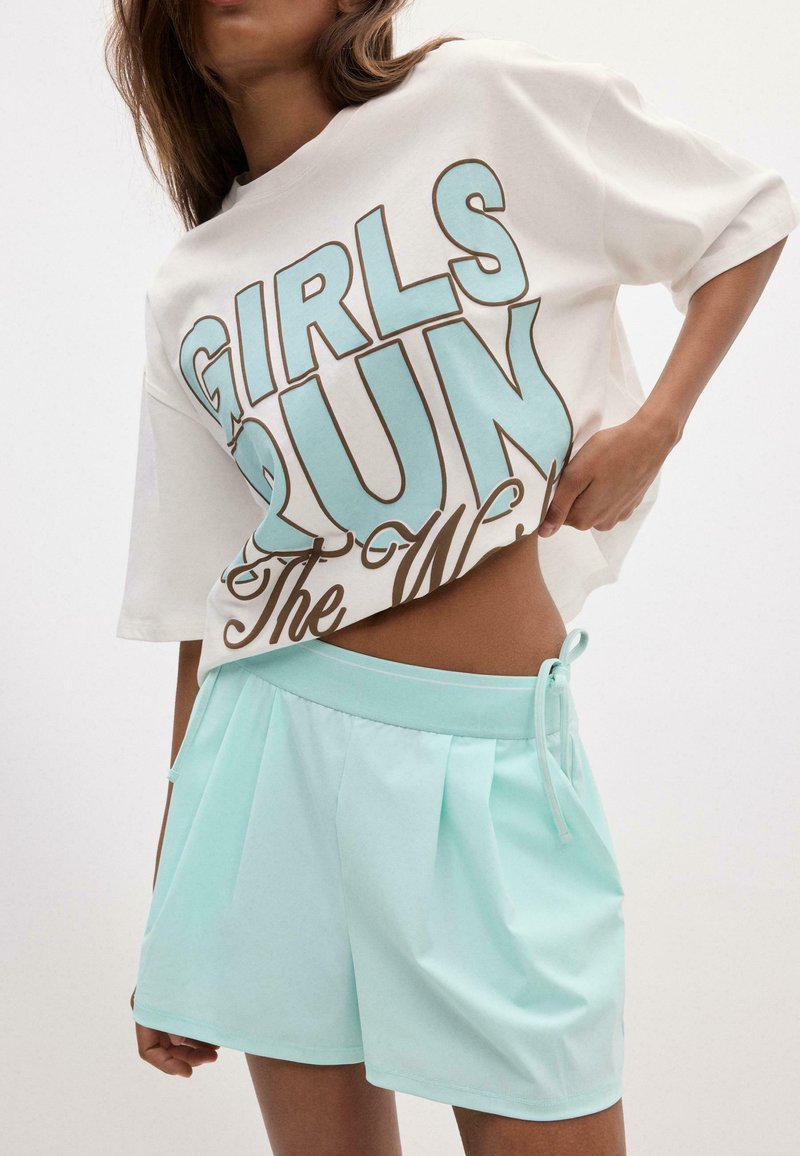 Femme portant un tee-shirt court blanc avec le texte "GIRLS RUN The World" et un short plissé bleu clair avec un nœud sur le côté.