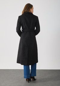 Black belted coat in textured fabric, knee-length, with a notched collar and long sleeves, paired with blue wide-leg jeans and black heels.