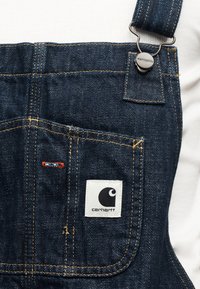 Κοντινό πλάνο στην τσέπη σκούρου μπλε τζιν φορμάκια με το λογότυπο Carhartt και μεταλλικό κούμπωμα στο λουράκι, πάνω σε λευκό ύφασμα φόντο.