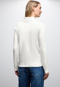 Witte langarmtop met hoge kraag, zachte textuur en rechte snit. Gecombineerd met een blauwe spijkerbroek met achterzakken.