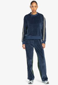 Set di tuta in velluto blu navy con collo a giro e polsini a coste, arricchito da strisce laterali con dettagli testuali. I pantaloni presentano pannelli verdi laterali.