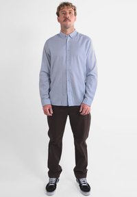 Chemise bleu clair à manches longues, associée à un pantalon marron foncé et des baskets noires. La chemise présente une texture subtile et un col.