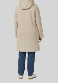 Beige lange jas met een gewatteerde capuchon, zachte textuur, rechte pasvorm en eenvoudig ontwerp. Gepaard met blauwe jeans en witte sneakers.