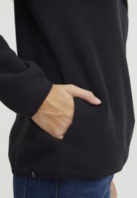 Sort fleece trøje med en tekstureret overflade, der har en sidelomme og tætsiddende manchetter, parret med blå denim jeans.