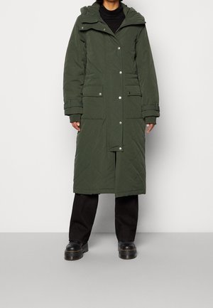 Personne portant un long manteau d'hiver matelassé vert foncé avec de grandes poches, un pantalon noir et des chaussures compensées noires, debout sur un sol gris.