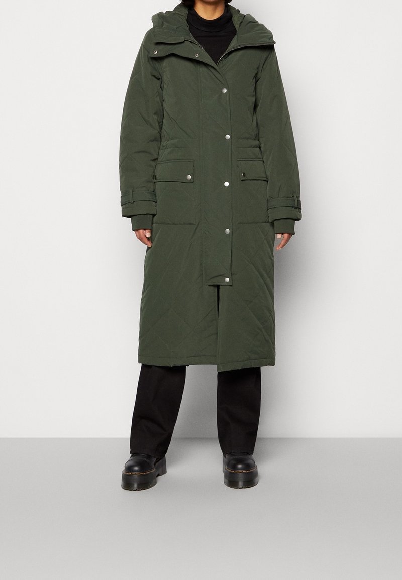 Personne portant un long manteau d'hiver matelassé vert foncé avec de grandes poches, un pantalon noir et des chaussures compensées noires, debout sur un sol gris.