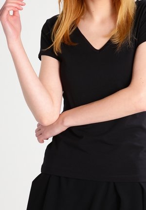 Femme à la peau claire et aux cheveux roux portant un t-shirt noir à col en V et manches courtes, tenant un bras avec l'autre sur un fond blanc.