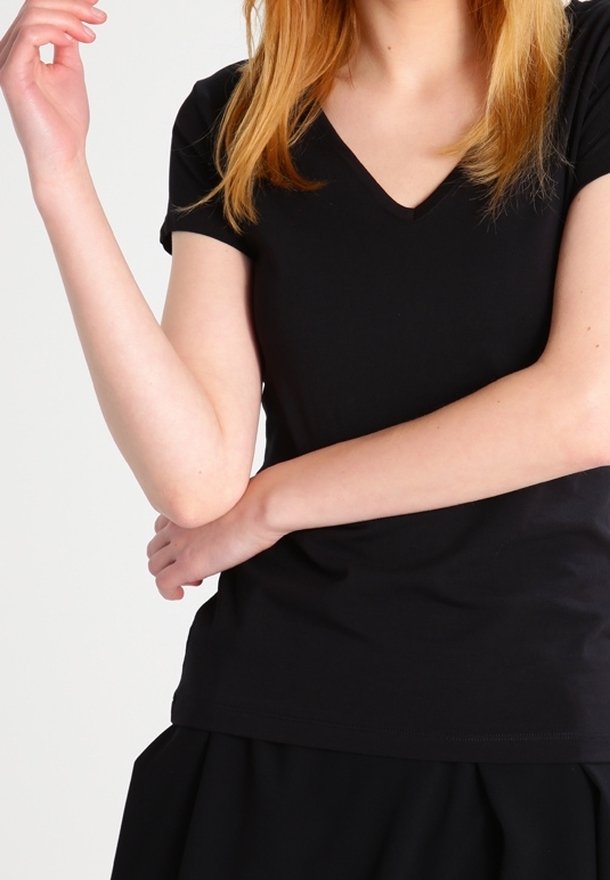 Femme à la peau claire et aux cheveux roux portant un t-shirt noir à col en V et manches courtes, tenant un bras avec l'autre sur un fond blanc.