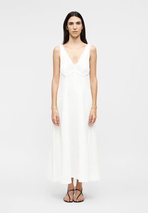 Kvinde med langt mørkt hår iført en ærmeløs hvid maxikjole med hæklede detaljer og sorte remsandaler, stående foran en ensfarvet baggrund.