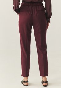 Pantalones burdeos con un corte relajado, cinturilla elástica, dos bolsillos traseros y diseño de pierna ajustada. La textura parece suave y lisa.