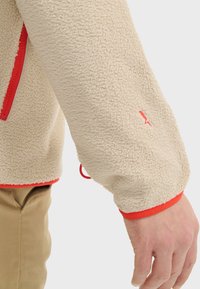 Veste en polaire beige avec une poche zippée rouge et des accents rouges aux poignets. Tissu texturé avec une finition lisse sur la manche.