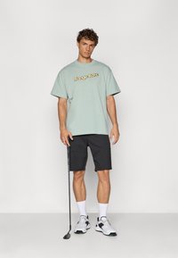 Lys grønn oversized t-skjorte med "Range Rats" grafikk, parret med svarte shorts, hvite sokker og joggesko. Golfkølle holdt i høyre hånd.