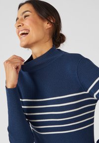 Maglione blu navy a costine con colletto alto, caratterizzato da strisce bianche orizzontali. Il materiale sembra morbido, con un design aderente.