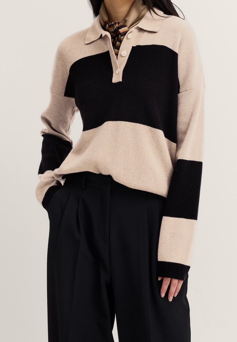 Femme portant un pull beige et noir à rayures manches longues avec col, associé à un pantalon noir plissé et un foulard rayé autour du cou.