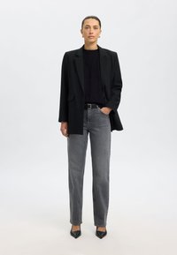 Črni blazer, oprijet črn top, sive kavbojke z visokim pasom in ravnimi nogavicami ter črne salonarji z zašiljenim nosom, na svetlo sivi podlagi.