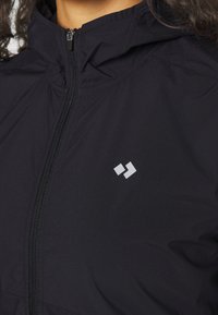 Černá zipová bunda s vysokým límcem, vyrobená z hladké, lehké tkaniny. Má malé bílé logo na levé straně.