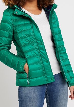 Nainen pukeutuneena teekkarin turkoosiin tikattuun takkiin valkoisen ribbineuleen ja sinisten farkkujen päällä, kädet takin taskuissa.