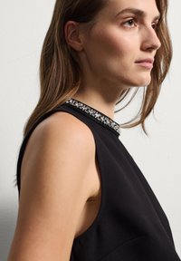 Vestido negro sin mangas con cuello alto que presenta un cuello adornado con cristales. Tejido suave y estructurado con una silueta ajustada.