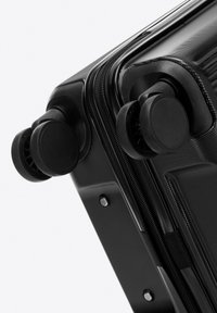 Zwarte harde koffer met een gestructureerde oppervlakte, dubbele zwarte wielen en ritsen. Heeft een slank ontwerp met afgeronde hoeken en minimale hardware.