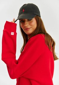 Čierna baseballová čiapka s logom, v kombinácii s červeným oversize mikinovým sveterom s rebrovanými manžetami a malou nášivkou. Model zakrýva ruku.