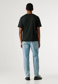 Μαύρη βαμβακερή μπλούζα με χαλαρή εφαρμογή, κοντά μανίκια, στρογγυλή λαιμόκοψη, που συνδυάζεται με ανοιχτό μπλε τζιν παντελόνι και μαύρα παπούτσια. Απλό φόντο.