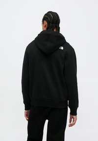 Osoba s pletenými vlasy, oblečená v černé mikině a černých kalhotách, otočená zády, s logem The North Face na horní části zad.