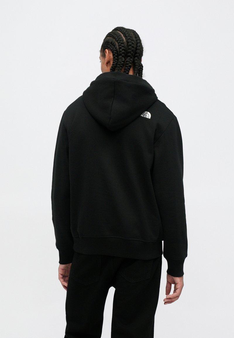 Personne avec des tresses portant un sweat à capuche noir et un pantalon noir, tourné vers l'arrière, avec le logo The North Face sur le haut du dos.