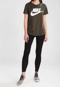 Olivgrön Nike-t-shirt med vitt logo, tillsammans med svarta leggings och grå löparskor med textilmaterial och svarta snörningar.