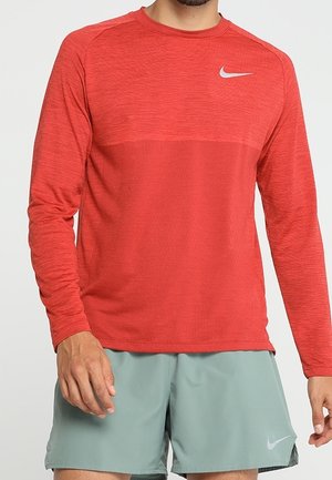 Mężczyzna w czerwonej, długorękawowej koszulce Nike i jasnoszarych sportowych szortach Nike, stojący z rękami swobodnie opuszczonymi wzdłuż ciała.