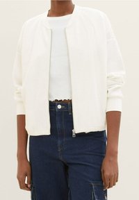 Giacca bomber bianca con texture a coste, zip frontale e polsini aderenti, indossata sopra un top bianco corto e abbinata a pantaloni di jeans scuri.