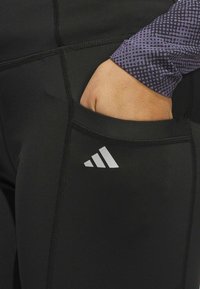 Zwarte sportleggings met een zijzak. De leggings hebben gestikte naden en een klein wit Adidas-logo nabij het onderbeen.