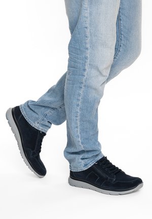 Person wearing light blue jeans and black sneakers with gray soles, standing with one leg crossed behind the other on a white background.