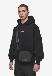 Čierna crossbody taška so zipsovými uzávermi, nastaviteľným pásom a embosovaným logom. Nosená cez čiernu mikinu a reflektujúce nohavice.