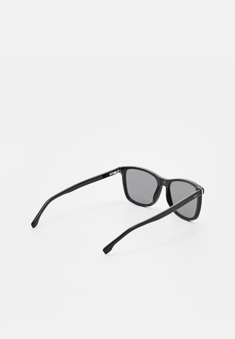 Schwarze rechteckige Sonnenbrille mit dunklen Gläsern, dünnen Bügeln und subtilen Markenlogo an dem rechten Schläfenbereich, auf einer weißen Oberfläche platziert.