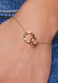 Złota bransoletka z dwoma połączonymi okrągłymi charmsami o teksturowanej powierzchni, spoczywająca na nadgarstku na tle dżinsu.