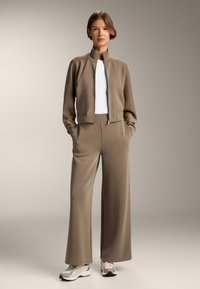 Beige rebrovaný skrátený kabát s vysokým golierom, skombinovaný s širokými nohavicami v rovnakom odtone. Model má na sebe biely top a tenisky. Celkový hladký povrch.