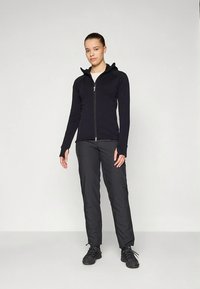 Sudadera negra con cremallera, cuello alto y mangas largas, combinada con pantalones negros de pierna recta y zapatos negros. Cuenta con ojal para el pulgar en los puños.