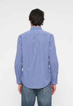 Homme portant une chemise à carreaux bleue et blanche à manches longues et un jean bleu, vu de dos sur un fond clair uni.