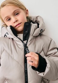 Giacca puffer grigia chiara con colletto alto, cuciture diagonali e dettagli neri. Presenta una chiusura con zip con logo sul cursore e sui polsini.