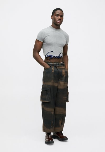 Grijze, aansluitende t-shirt met blauw logo op de taille, gecombineerd met losse zwarte cargobroeken met bruine tie-dye patronen en meerdere zakken. Bruine veterschoenen.