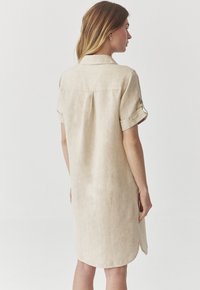 Vestito camicia in lino beige con maniche corte, colletto a bottoni e piega sul retro, caratterizzato da un orlo arrotondato e una texture liscia.
