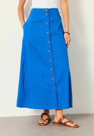 Frau trägt einen leuchtend blauen Knopfvorder-Midirock mit Taschen und braunen flachen Sandalen, steht auf einem grauen Boden.