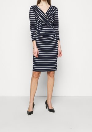 Frau trägt ein knielanges dunkelblaues Kleid mit weißen horizontalen Streifen und dreiviertel Ärmeln, kombiniert mit schwarzen High Heels.