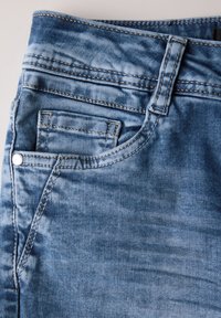 Tela de mezclilla azul con un aspecto desgastado. Cuenta con un bolsillo frontal, costuras visibles y un detalle de botón de metal en la cinturilla.