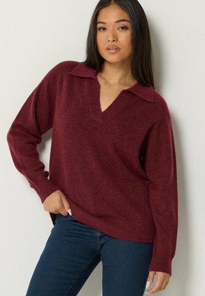 Frau mit langen dunklen Haaren trägt einen lockeren maroonfarbenen Strickpullover mit Kragen und dunkelblaue Jeans und steht vor einer hellen Wand.